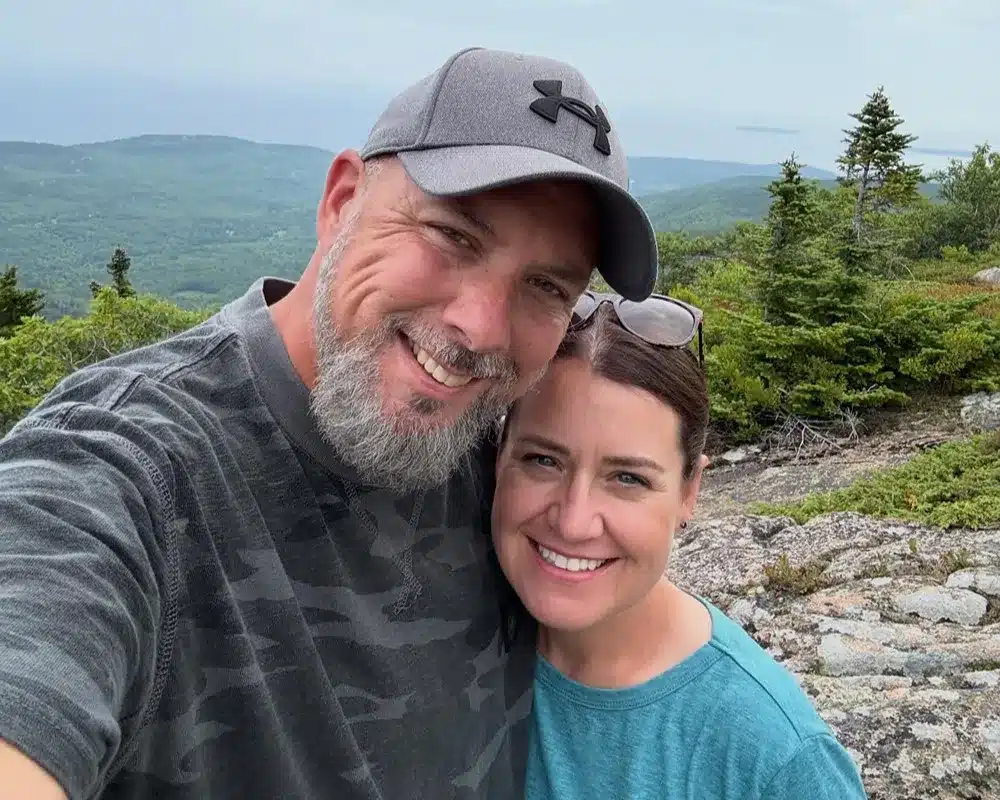 Smiling couple taking a selfie on a rocky mountaintop with trees and distant hills behind them.