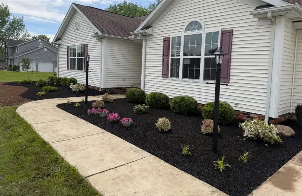 White suburban house with arched window and two black lamp posts, front yard landscaped with mulch, trimmed shrubs, and a curved concrete walkway extending from the lawn to the entrance.